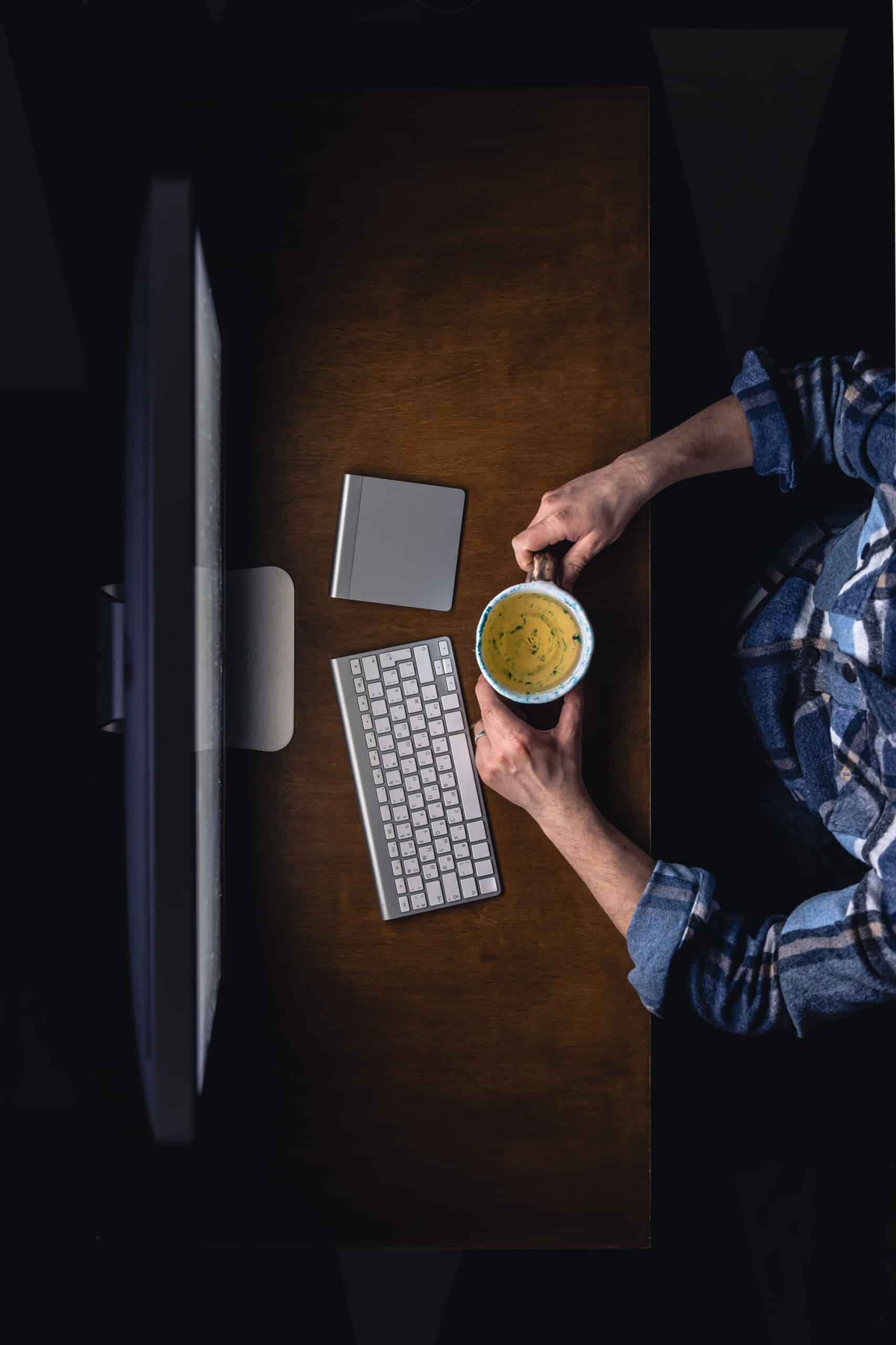 top view of man working on computer at night, holding mug of tea.